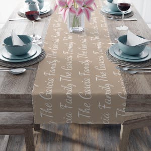 Puede incluir: Un camino de mesa beige con el texto "The Garcia Family" en escritura blanca. El camino de mesa está sobre una mesa de madera preparada para una comida, con platos, cuencos, cubiertos y copas de vino. Un jarrón con flores rosas está en el centro.