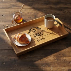 Puede incluir: Una bandeja rectangular de bambú con asas, que contiene una taza blanca, un plato con un cruasán y un tarro de miel con un sumergidor. La bandeja está grabada con el texto "A JONES family".