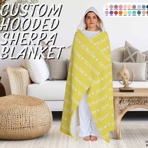 K&ouml;nnte beinhalten: Eine gelbe Sherpa-Decke mit Kapuze mit wei&szlig;en Herzen und den Namen "Evelyn & Ethan", die sich durchgehend wiederholen. Die Decke ist &uuml;ber ein Sofa gelegt und eine Person tr&auml;gt die Decke als Kapuzenrobe.