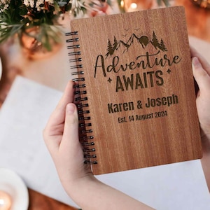 May include: A wooden journal with a spiral binding. The cover is engraved with a mountain scene and the text "Adventure Awaits". The names "Karen & Joseph" and the date "Est. 14 August 2024" are also engraved on the cover.