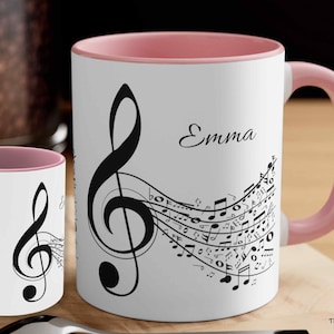 May include: A white ceramic mug with a pink rim and handle. The mug features a black treble clef and musical notes with the name "Emma" printed on the front.