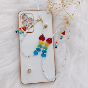 May include: A white phone case with a rainbow beaded phone strap and a small fish charm. The strap is attached to the case with a gold ring.