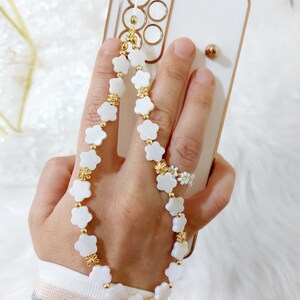Puede incluir: Funda de teléfono blanca con una correa de teléfono de flores blancas y doradas. La correa está hecha de pequeñas cuentas de flores blancas con detalles dorados.