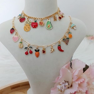 May include: Two gold necklaces with colourful fruit charms, including strawberries, pineapples, apples, and avocados. The necklaces are displayed on a white mannequin bust. A pink flower is in the foreground.