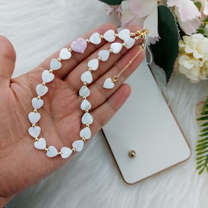 Può includere: Una collana di perline a forma di cuore bianche e dorate con un ciondolo a forma di cuore rosa. La collana è su una mano e ha una catena d'oro con un ciondolo a forma di cuore.