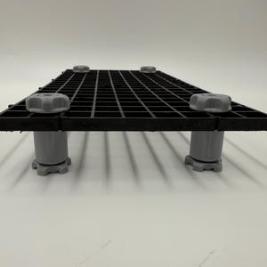 May include: A black plastic grid with a square pattern, supported by four gray adjustable legs. Each leg has a gray knob on top. The grid is designed to elevate items.