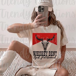 Könnte beinhalten: Ein weißes T-Shirt mit einer roten und weißen Grafik eines Bullenschädels mit dem Text "Whiskey Bent and Hellbound". Das Model trägt eine beige Baseballkappe mit dem Wort "Rebel" darauf.