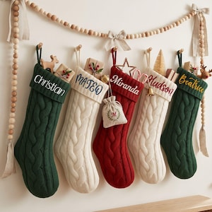 May include: Five cable-knit Christmas stockings in dark green, cream, and red, each featuring a personalised name embroidered on the cuff. The stockings are hung from a wooden bead garland and contain small gifts, ornaments, and a tiny pouch.