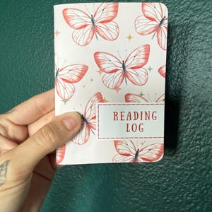 May include: A small notebook with a white cover featuring a repeating pattern of orange butterflies and small gold stars. A rectangular white label with a red dashed border is printed on the front with the red text READING LOG.