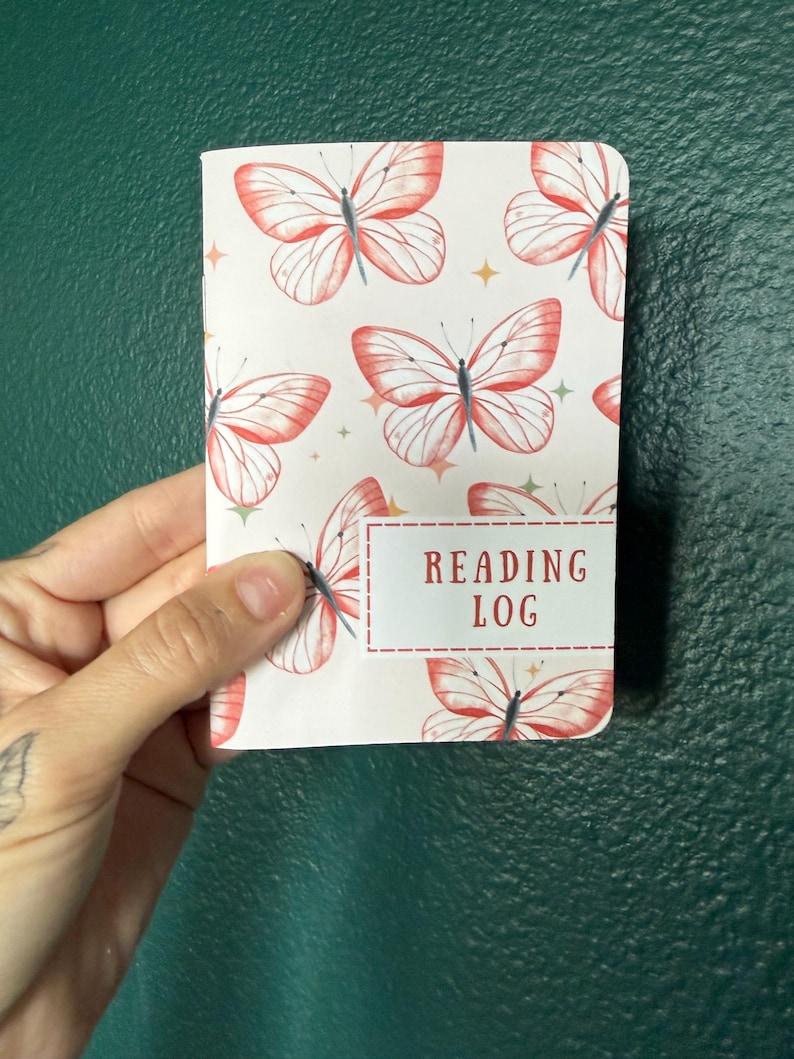 May include: A small notebook with a white cover featuring a repeating pattern of orange butterflies and small gold stars. A rectangular white label with a red dashed border is printed on the front with the red text READING LOG.
