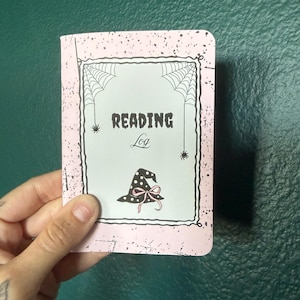 May include: A rectangular reading log notebook with a light pink cover featuring black speckles. The center has a white rectangular frame with spiderwebs in the corners, two small spiders, and a black witch hat with a pink bow. Text reads READING Log.