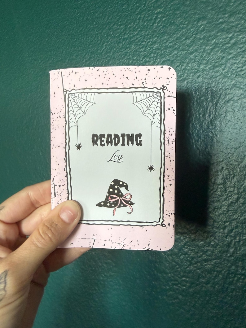May include: A rectangular reading log notebook with a light pink cover featuring black speckles. The center has a white rectangular frame with spiderwebs in the corners, two small spiders, and a black witch hat with a pink bow. Text reads READING Log.