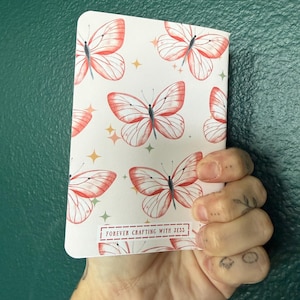 May include: A rectangular notebook with a white cover featuring a repeating pattern of red-orange butterflies and small yellow four-pointed stars. The bottom center of the cover displays a rectangular label with the text FOREVER CRAFTING WITH JESS.