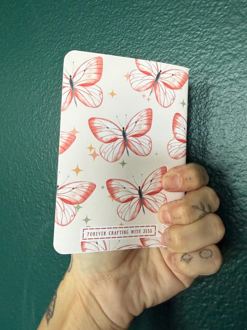 May include: A rectangular notebook with a white cover featuring a repeating pattern of red-orange butterflies and small yellow four-pointed stars. The bottom center of the cover displays a rectangular label with the text FOREVER CRAFTING WITH JESS.