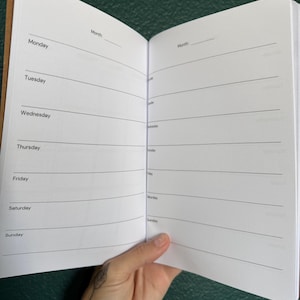 May include: An open planner with white pages featuring black text and horizontal lines. The left page lists the days of the week from Monday to Sunday, while the right page provides blank lines for notes. Both pages include a space for the month.