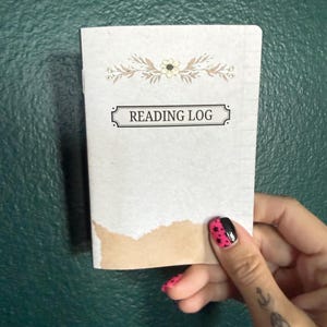 May include: A small rectangular reading log notebook with a light gray cover featuring a floral illustration at the top and the text READING LOG centered in a black rectangular frame. The bottom edge of the cover has a torn brown paper accent.