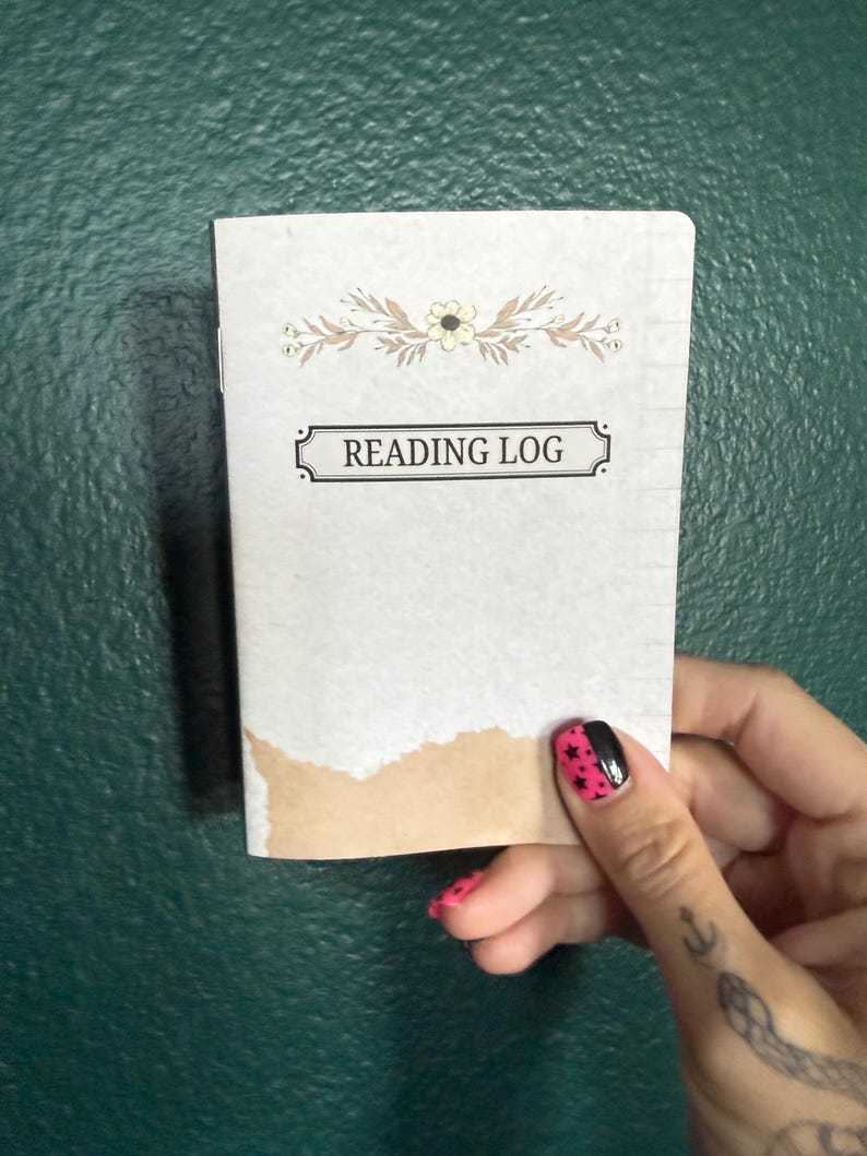 May include: A small rectangular reading log notebook with a light gray cover featuring a floral illustration at the top and the text READING LOG centered in a black rectangular frame. The bottom edge of the cover has a torn brown paper accent.