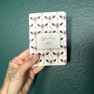 May include: A rectangular reading log notebook with a white cover featuring a repeating pattern of pink eyes with black bat wings and small pink teardrops. The center of the cover has a white rectangular label with the text Reading Log in black.