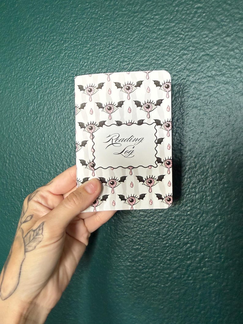 May include: A rectangular reading log notebook with a white cover featuring a repeating pattern of pink eyes with black bat wings and small pink teardrops. The center of the cover has a white rectangular label with the text Reading Log in black.