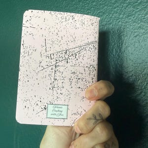 May include: A rectangular notebook with a light pink cover featuring a black distressed, speckled pattern. At the bottom center, a small rectangular label displays the text Forever Crafting with Jess in a decorative script font.