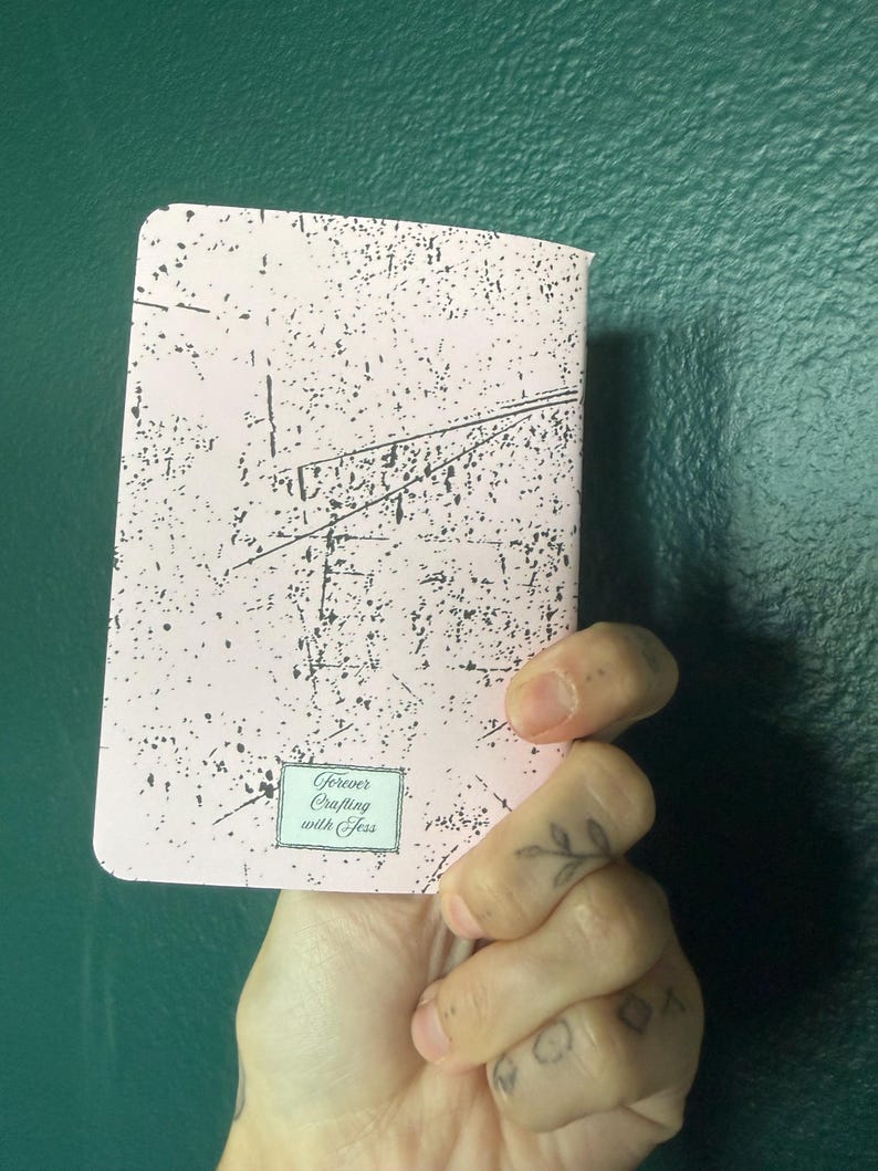 May include: A rectangular notebook with a light pink cover featuring a black distressed, speckled pattern. At the bottom center, a small rectangular label displays the text Forever Crafting with Jess in a decorative script font.