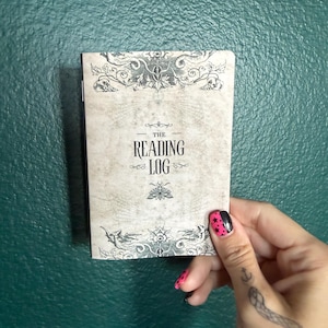 May include: A rectangular notebook with a beige cover featuring the text THE READING LOG in black lettering. The cover design includes intricate black illustrations of floral patterns, a moth, and dragon-like creatures at the top and bottom edges.