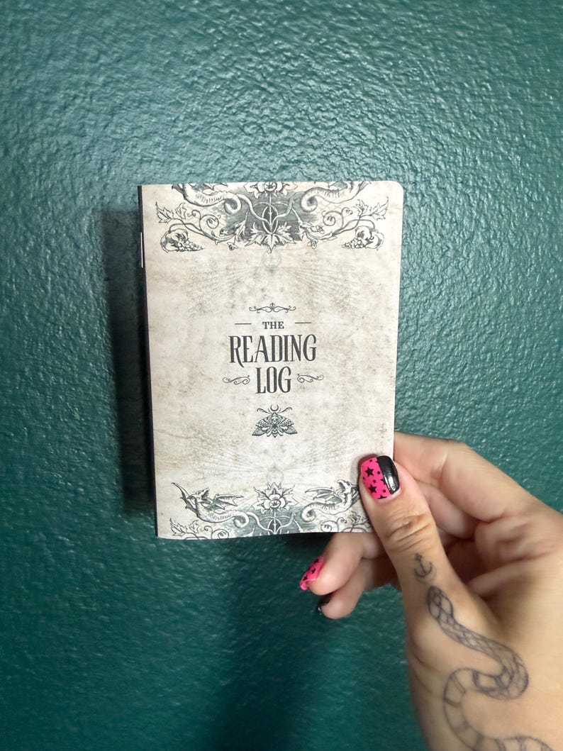 May include: A rectangular notebook with a beige cover featuring the text THE READING LOG in black lettering. The cover design includes intricate black illustrations of floral patterns, a moth, and dragon-like creatures at the top and bottom edges.