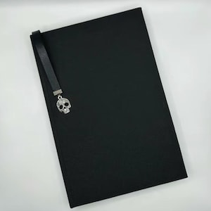 May include: A black journal with a matching black bookmark. The bookmark features a silver skull charm with crystal accents. The journal is rectangular and appears to be made of a textured material.