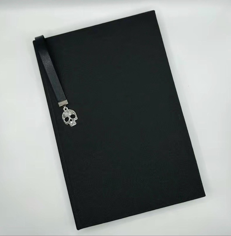 May include: A black journal with a matching black bookmark. The bookmark features a silver skull charm with crystal accents. The journal is rectangular and appears to be made of a textured material.