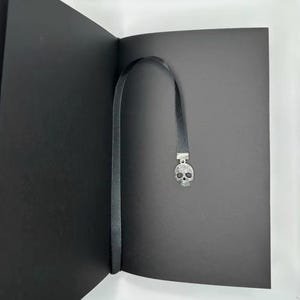 May include: A black bookmark with a silver skull charm is inserted into a black book. The bookmark is a thin, black ribbon. The skull charm is detailed with small, sparkling accents, adding a touch of gothic flair to the reading accessory.