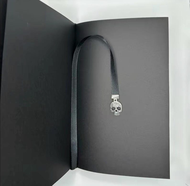 May include: A black bookmark with a silver skull charm is inserted into a black book. The bookmark is a thin, black ribbon. The skull charm is detailed with small, sparkling accents, adding a touch of gothic flair to the reading accessory.