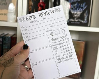 May include: A white book review sheet with the title "BOOK REVIEW" at the top. The sheet includes sections for title, author, genre, and a rating system. The sheet is held in a person's hand.