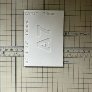 May include: A white rectangular A7 paper size template with engraved text including 2.913 inches, 4.134 inches, 74mm, and 105mm. The template is positioned on a grid mat with a ruler showing measurements in inches and centimeters.