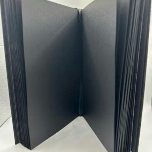 May include: An open, black photo album with thick, black pages. The album is bound with a black spine and is fanned open, revealing the pages. The album is designed for photographs and keepsakes.