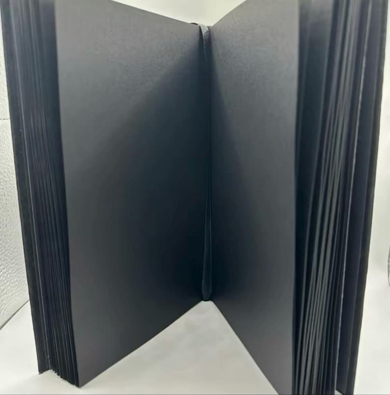 May include: An open, black photo album with thick, black pages. The album is bound with a black spine and is fanned open, revealing the pages. The album is designed for photographs and keepsakes.
