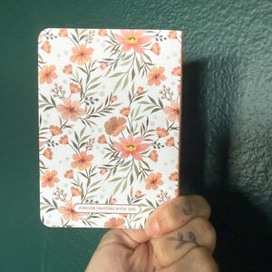 May include: A rectangular notebook featuring a white cover with a repeating pattern of peach-colored flowers, green leaves, and small gray dots. A small rectangular label at the bottom center contains the text FOREVER CRAFTING WITH JESS.