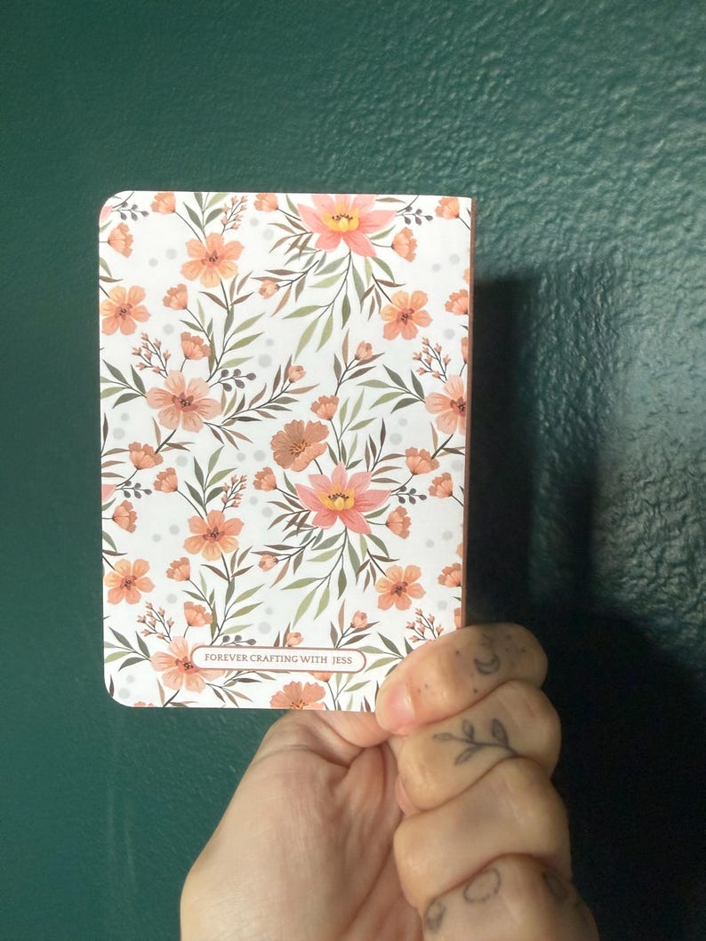 May include: A rectangular notebook featuring a white cover with a repeating pattern of peach-colored flowers, green leaves, and small gray dots. A small rectangular label at the bottom center contains the text FOREVER CRAFTING WITH JESS.