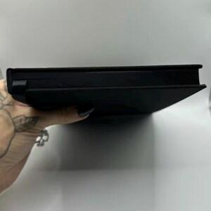 May include: A black book with a textured cover and a black ribbon bookmark. The book is closed and appears to be new. The spine is visible, and the pages are neatly bound.