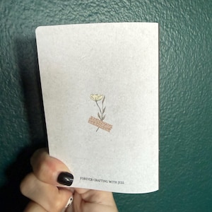 May include: A white rectangular card featuring a small illustration of a single yellow flower with green leaves, accented by a piece of tan patterned tape at the base. The text FOREVER CRAFTING WITH JESS is printed in black at the bottom center.