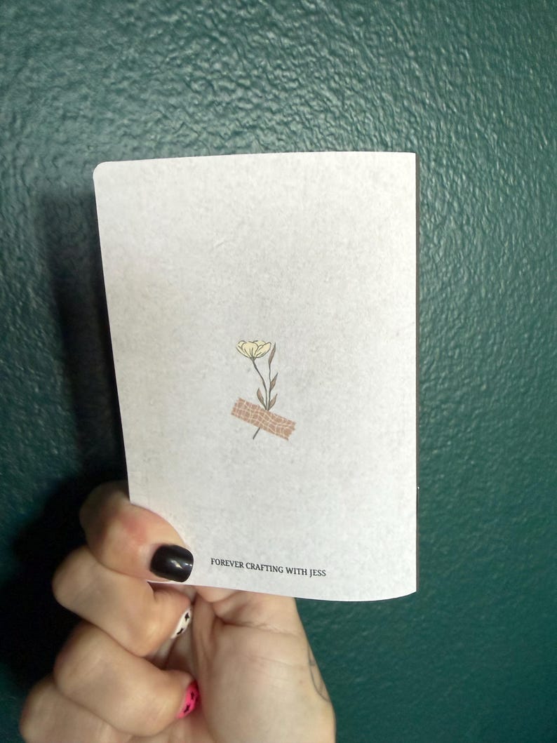 May include: A white rectangular card featuring a small illustration of a single yellow flower with green leaves, accented by a piece of tan patterned tape at the base. The text FOREVER CRAFTING WITH JESS is printed in black at the bottom center.