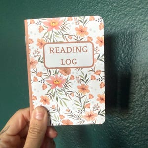 May include: A rectangular reading log notebook featuring a white cover with a repeating pattern of peach-colored flowers and green foliage. The center of the cover displays a rectangular box with the text READING LOG printed in dark brown letters.