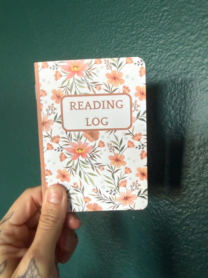 May include: A rectangular reading log notebook featuring a white cover with a repeating pattern of peach-colored flowers and green foliage. The center of the cover displays a rectangular box with the text READING LOG printed in dark brown letters.