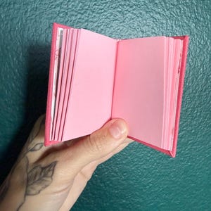 May include: A miniature open book with a bright pink fabric cover and numerous blank light pink pages. The binding is visible at the center, and the book is held open to display the stack of paper leaves on both the left and right sides.