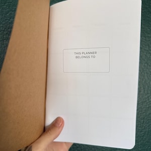 May include: A planner with a brown paper cover and white interior pages. The first page features a rectangular box with the printed text THIS PLANNER BELONGS TO centered inside. The planner has a simple design with a grid layout visible on the page.