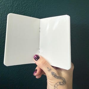 May include: A small, open, blank white booklet with rounded corners. The booklet features a simple center-fold binding with visible staples along the spine. The pages are plain, smooth, and white, providing a clean surface for writing or drawing.