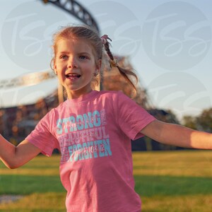 May include: A pink t-shirt with the words "STRONG, BEAUTIFUL, FORGIVEN" in blue and white text. The shirt also has the text "Philippians 4:13" and "Psalm 103:12". The shirt is being worn by a young girl.