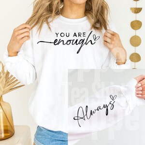 May include: White sweatshirt with the words "YOU ARE enough" in black script across the chest. The sleeve features the word "Always" in black script. The sweatshirt is a long-sleeved, crew neck style.