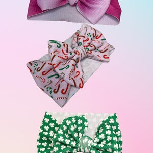 May include: Three fabric headbands with different bow designs. The first headband has a pink ombre bow, the second headband has a white bow with a red and green candy cane pattern, and the third headband has a green bow with a white clover pattern.