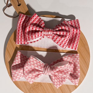 May include: Two red and white patterned hair bows. One bow has a red and white gingham pattern. The other bow has a red and white pattern with small hearts.