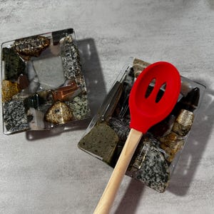 May include: Two clear glass spoon rests with a mosaic of colorful stones. A red silicone slotted spoon with a wooden handle is resting on one of the spoon rests.